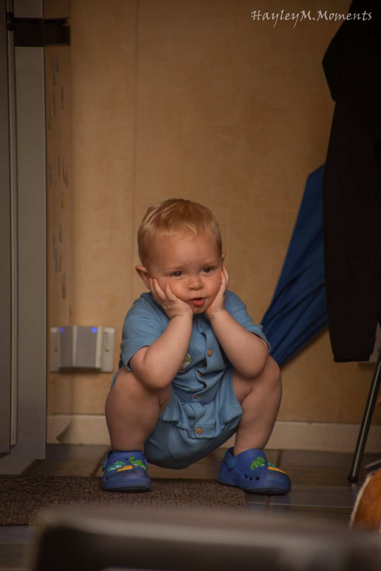 Toddler squatting in a hallway with his hands on his cheeks